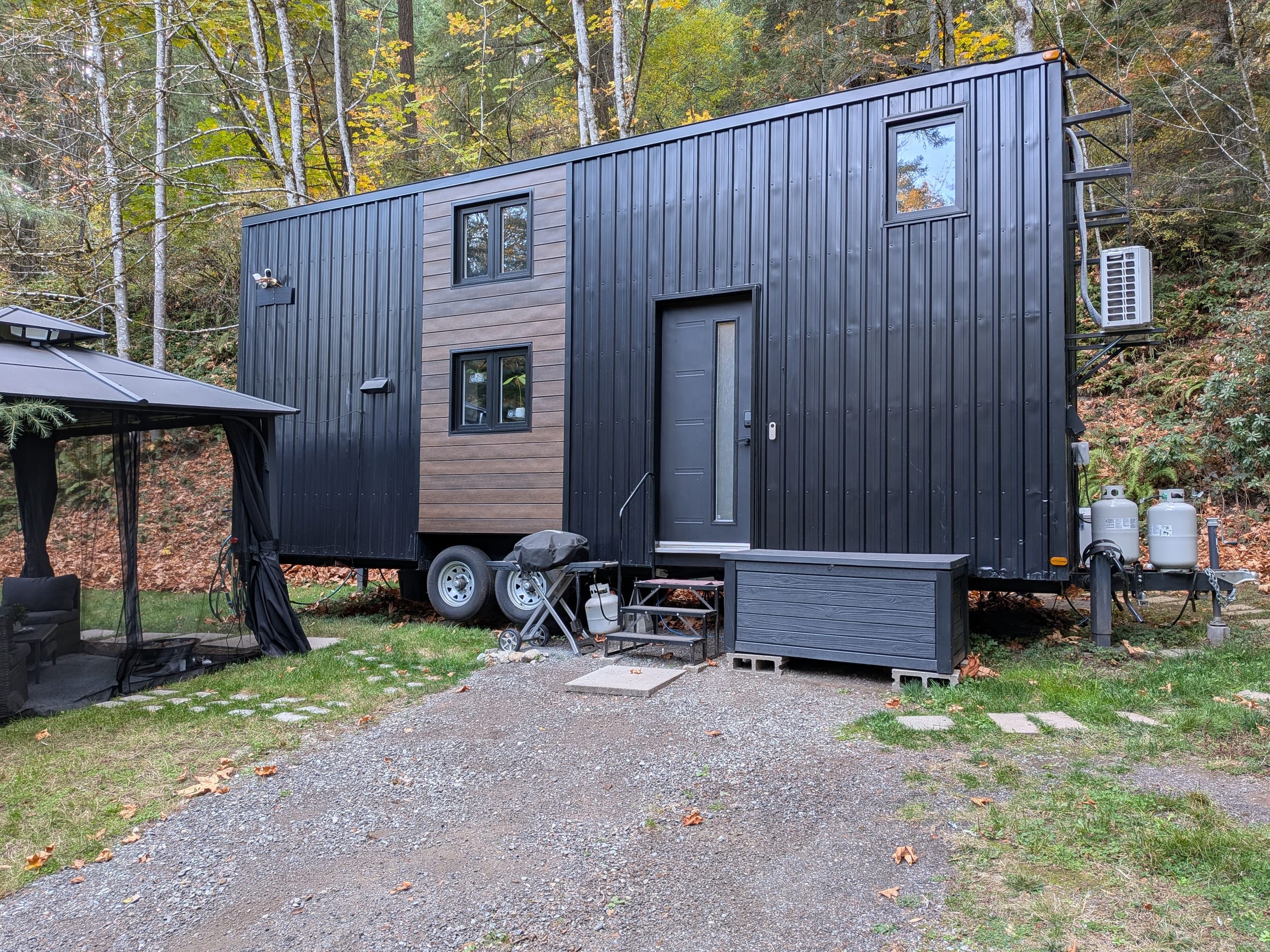Steel-clad tiny house on wheels with wood accent siding, front door, gazebo and gravel driveway