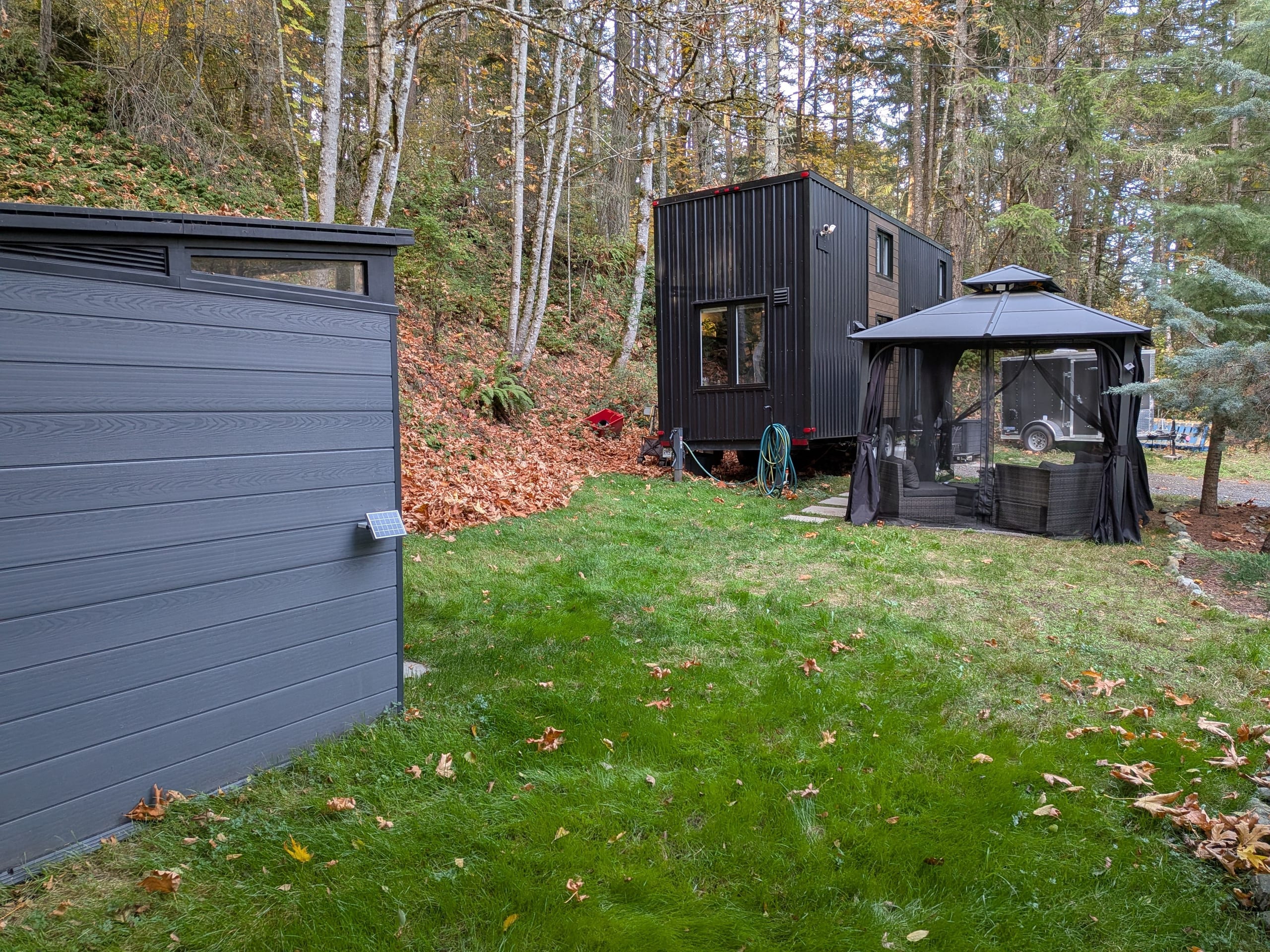 Two grey storage sheds on the lawn with the tiny house and gazebo beyond